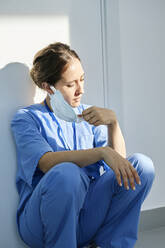 Nurse removing protective face mask squatting by wall in hospital - DSHF00532