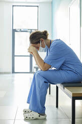 Exhausted nurse wearing protective face mask sitting on bench in corridor - DSHF00544