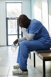Müder Arzt mit dem Kopf in der Hand auf einer Bank im Krankenhauskorridor sitzend - DSHF00545