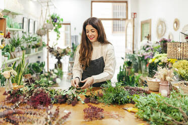 Smiling woman arranging leaves on workbench at floral store - MRRF02407