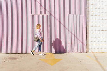 Woman walking in front of corrugated wall on sunny day - MGRF00744