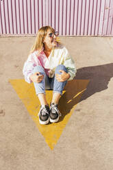 Happy young woman sitting on arrow symbol - MGRF00748