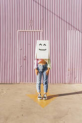 Frau, die eine Schachtel mit Robotergesicht trägt, steht vor einer Wellblechwand - MGRF00751