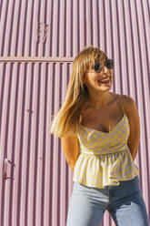 Happy woman wearing sunglasses standing in front of corrugated wall - MGRF00755