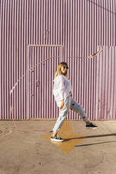 Woman playing with rainbow colored gymnastics ribbon in front of corrugated wall - MGRF00756