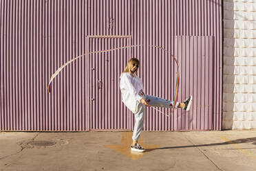 Frau steht auf einem Bein und spielt mit einem regenbogenfarbenen Gymnastikband vor einer Wellblechwand - MGRF00757