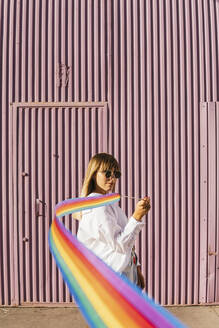 Frau hält regenbogenfarbenes Gymnastikband vor einer Wellblechwand - MGRF00758