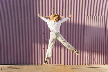 Cheerful woman jumping with arms outstretched in front of corrugated wall - MGRF00774