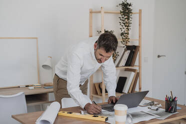 Älterer Architekt mit Blaupause und Laptop bei der Arbeit am Schreibtisch - EBBF06078