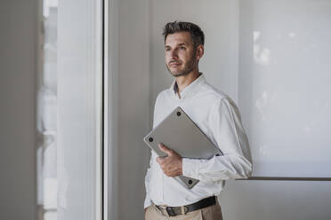 Thoughtful businessman holding laptop looking through window standing with hand in pocket at office - EBBF06084