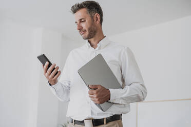 Smiling businessman holding laptop looking at mobile phone in office - EBBF06087