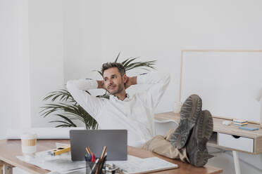 Smiling businessman with hands behind head resting on chair in office - EBBF06089