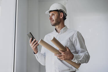 Engineer holding rolled up paper and smart phone in front of white wall - EBBF06093