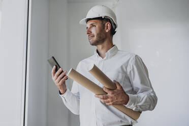 Ingenieur hält aufgerolltes Papier und Smartphone vor einer weißen Wand - EBBF06093