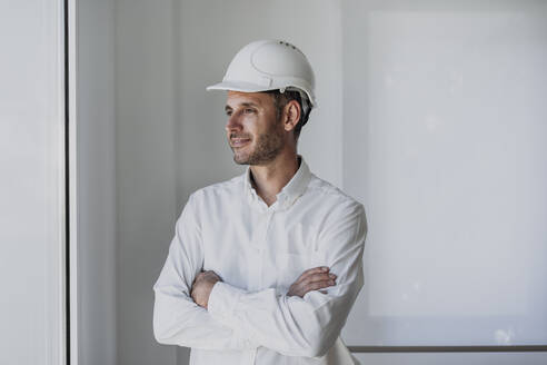 Lächelnder Ingenieur mit verschränkten Armen, der durch ein Fenster vor einer weißen Wand schaut - EBBF06094