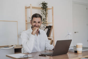 Smiling businessman sitting with laptop at desk in office - EBBF06113