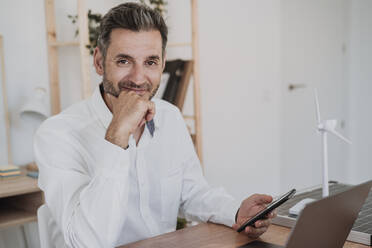 Smiling businessman holding smart phone sitting with laptop at desk - EBBF06114