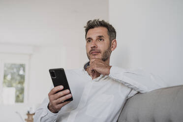 Nachdenklicher Geschäftsmann mit Smartphone auf dem Sofa im Büro - EBBF06131