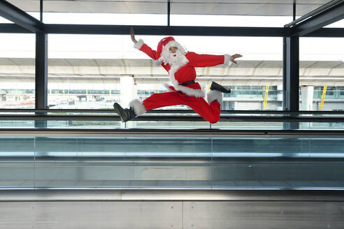 Man wearing Santa Claus costume jumping on moving walkway at airport - GGGF00953