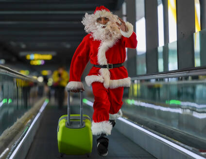 Man wearing Santa Claus costume running with suitcase on moving walkway at airport - GGGF00954