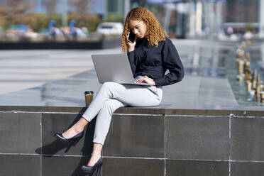 Young freelancer using laptop and talking on smart phone - GGGF00960