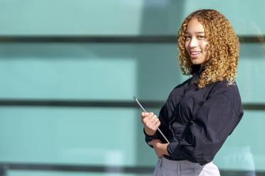 Lächelnde junge Geschäftsfrau mit blondem Lockenkopf, die an einem sonnigen Tag einen Tablet-PC hält - GGGF00969