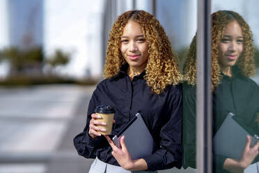 Smiling young businesswoman holding coffee cup leaning on glass - GGGF00974