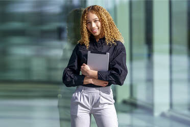 Happy young businesswoman with curly hair standing in front of glass - GGGF00978