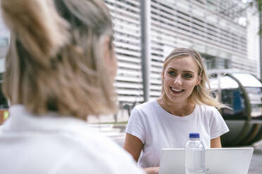 Happy businesswoman with laptop talking to colleague at office park - AMWF00465