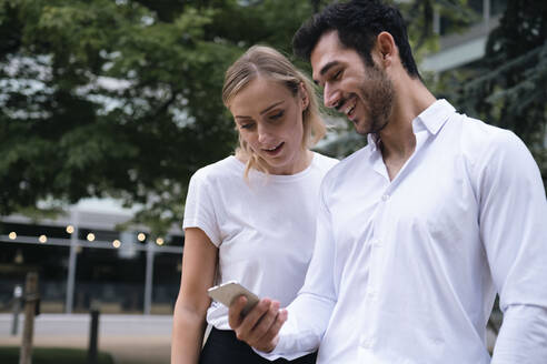 Glücklicher junger Geschäftsmann, der sein Smartphone mit einem Kollegen im Büropark benutzt - AMWF00485