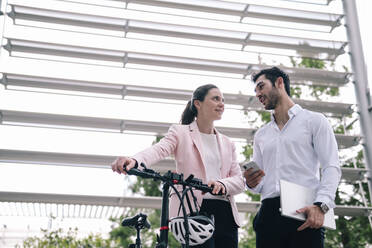 Lächelnde Geschäftsfrau mit Fahrrad im Gespräch mit einem Kollegen im Büropark - AMWF00487