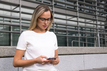 Businesswoman using smart phone sitting in front of building at office park - AMWF00501