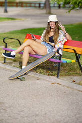 Happy woman showing peace gesture sitting on colorful park bench - GGGF00982