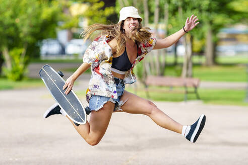 Carefree woman with skateboard jumping at park - GGGF00985