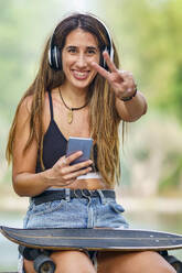 Glückliche junge Frau mit Smartphone und Friedenszeichen-Geste - GGGF00986