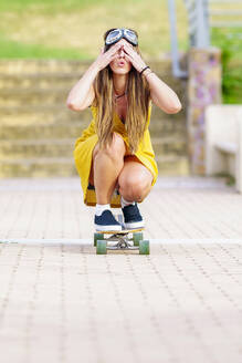 Junge Frau, die mit der Hand die Augen bedeckt und auf dem Gehweg Skateboard fährt - GGGF00990