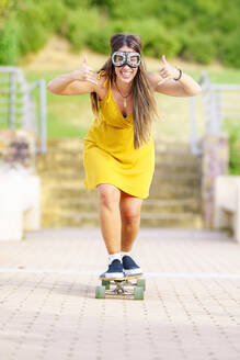 Glückliche junge Frau mit Shaka-Zeichen und Skateboard auf dem Fußweg - GGGF00992