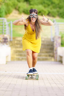 Happy young woman gesturing Shaka sign and skateboarding on footpath - GGGF00992