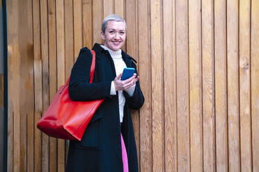Happy woman with mobile phone leaning on wooden wall - AMWF00513