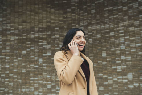 Glückliche junge Frau, die vor einer Wand mit einem Smartphone spricht - AMWF00519