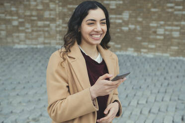 Glückliche junge Frau mit Smartphone an der Wand stehend - AMWF00520