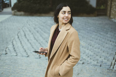 Smiling young woman in trench coat holding smart phone - AMWF00527