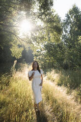 Woman wearing white dress standing in forest on sunny day - EYAF02108
