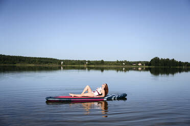 Frau entspannt auf SUP-Board im See unter blauem Himmel - EYAF02112