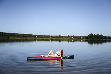 Woman relaxing on SUP board in lake under blue sky - EYAF02112