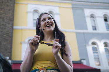 Happy woman holding sunglasses day dreaming in front of building - AMWF00552