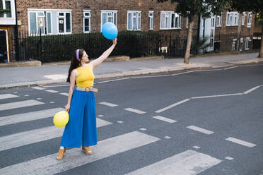 Frau mit erhobener Hand und Luftballons auf dem Zebrastreifen stehend - AMWF00569