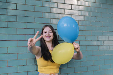 Woman holding blue and yellow balloons showing peace sign in front of wall - AMWF00570