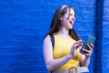 Cheerful woman holding mobile phone in front of blue wall - AMWF00575