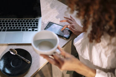 Hände einer Geschäftsfrau, die eine Kaffeetasse hält und ein Smartphone benutzt, während sie mit einem Laptop am Schreibtisch sitzt - AMWF00583